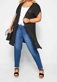 Svart lång cardigan med öppen framsida och korta ärmar, kombinerad med en vit knapp-up topp och blå tajta jeans. Svart handväska och klacksandaler.