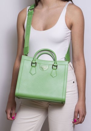 Frau trägt weißes Tanktop und Hose, trägt grüne strukturierte Handtasche mit doppelten Griffen und breitem Schulterriemen quer über der Brust.