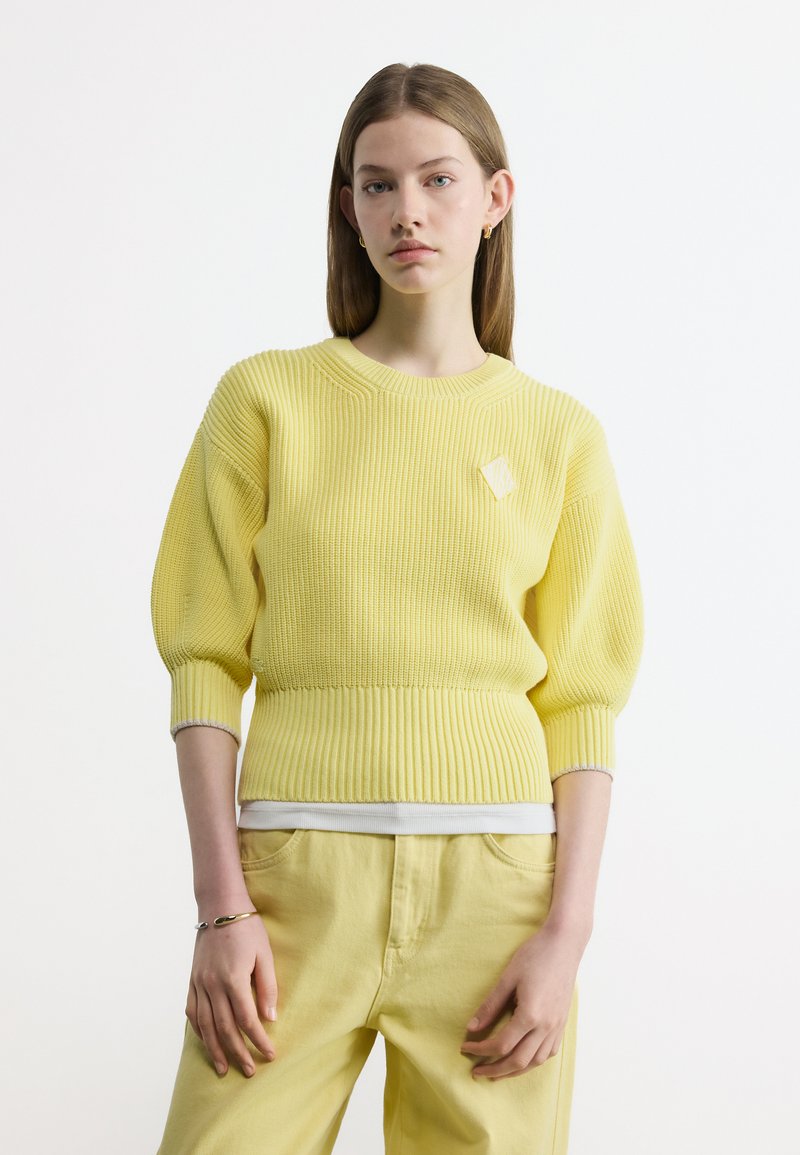Νεαρή γυναίκα με ίσια μαλλιά φοράει κίτρινο πουλόβερ με ριπ υφή και φουσκωτά μανίκια και κίτρινο παντελόνι, στέκεται μπροστά σε λευκό φόντο.