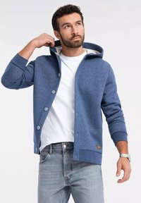 Sudadera azul texturizada con botones, un forro interior en contraste y un bolsillo frontal; combinada con una camiseta blanca y jeans claros.