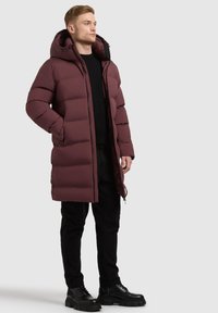 Burgundy puffer coat with a hood, featuring a zip closure, quilted design, and a knee-length cut, paired with black pants and boots.