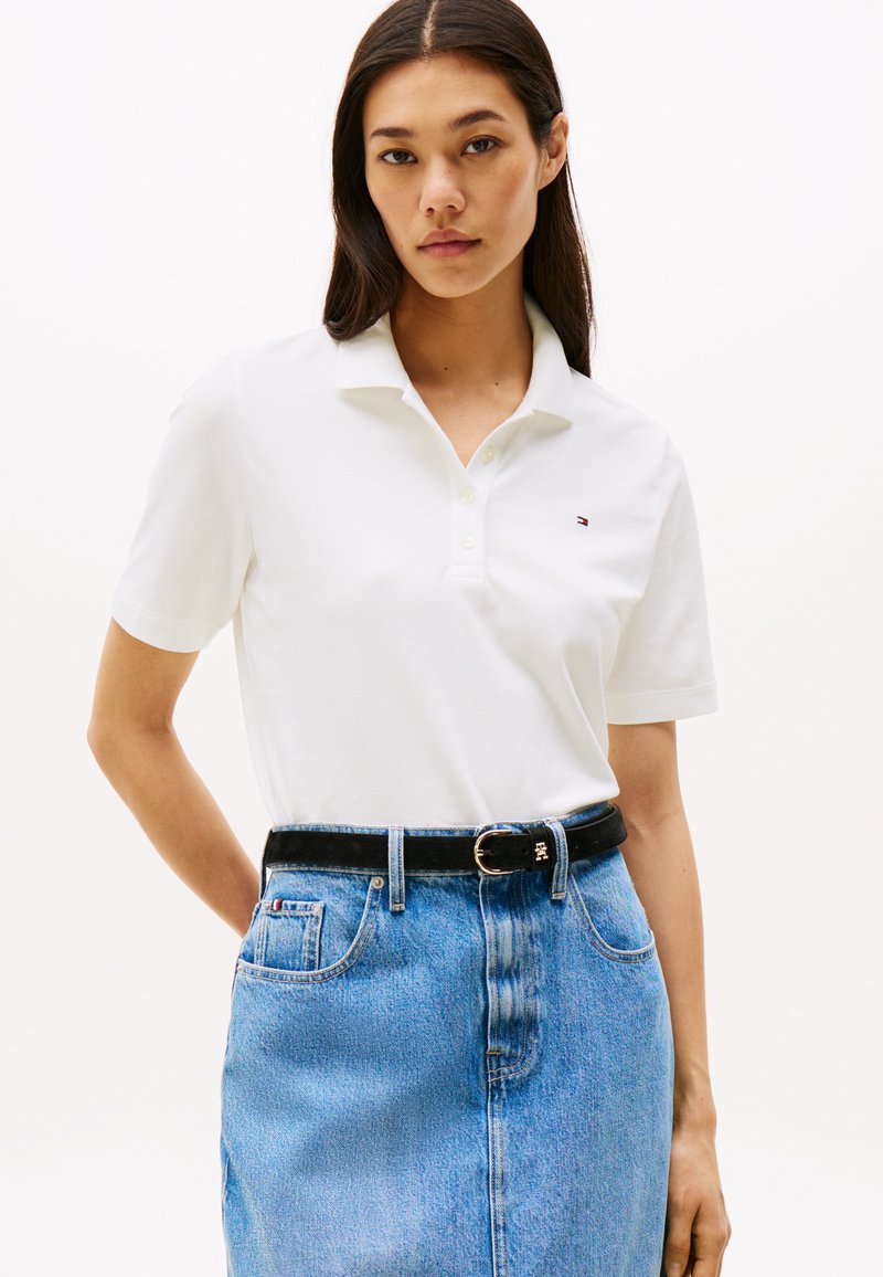 Young woman with long dark hair wearing a white polo shirt tucked into a high-waisted blue denim skirt with a black belt.