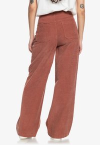 Pantalons en velours côtelé amples de couleur rouille, à taille haute, avec une texture à côtes verticales et deux grandes poches arrière.