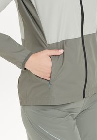 Lichtgewicht, tweekleurige jas met een ritsvak in olijfgroen en grijs. Voorzien van elastische manchetten en een ritssluiting aan de voorkant met zwarte afwerking.