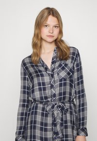 Camicia a quadri blu navy e bianca con maniche lunghe, chiusura con bottoni frontale, taschino sul petto e cintura in vita. Il tessuto sembra morbido e leggero.