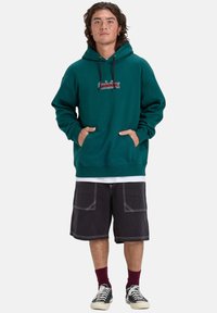 Sudadera color verde azulado con un bolsillo frontal y cordones. Presenta un bordado de logo en rojo y blanco. Combinada con pantalones cortos negros y zapatillas a rayas.