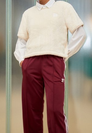 Persoon die een wit overhemd met kraag draagt onder een crèmekleurige, pluizige korte mouwtrui, gecombineerd met bordeauxrode Adidas trainingsbroek met witte strepen.