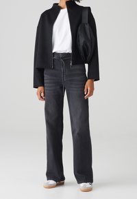 Chaqueta negra corta con cuello alto, camiseta blanca debajo, pantalones vaqueros de pierna ancha de mezclilla oscura y zapatillas blancas con suelas de goma.