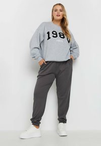 Grijze sweatshirt met zwarte "1984 PARIS" print, gecombineerd met donkergrijze geribbelde joggers. Beide hebben een ontspannen pasvorm. Witte sneakers maken de look af.