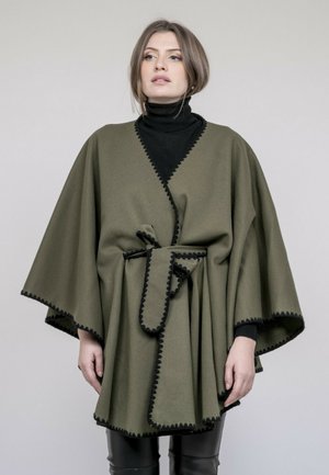 Femme portant une cape vert olive ceinturée avec un liseré noir par-dessus un col roulé noir, debout devant un fond uni et clair.