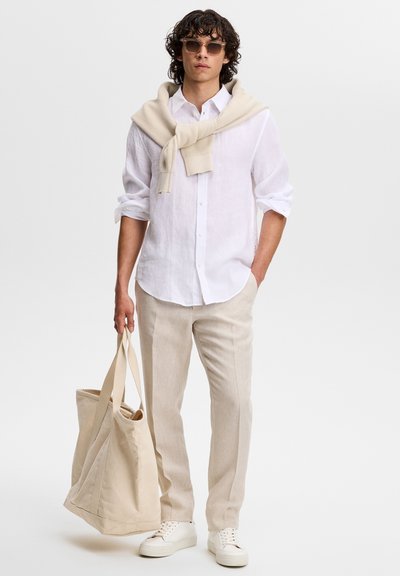 Jeune homme portant des lunettes de soleil, une chemise blanche, un pantalon beige, un pull noué sur les épaules, tenant un grand sac fourre-tout beige, debout devant un fond blanc.