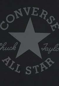 Čierna tkanina s veľkou sivou hviezdou v strede, obklopenou nápismi "CONVERSE ALL STAR" a "Chuck Taylor" v písanom písme.