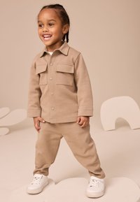 Ensemble pour enfant beige en deux pièces, en coton. La veste comporte deux poches avant et des boutons-pression; le pantalon a une coupe décontractée. Baskets blanches.