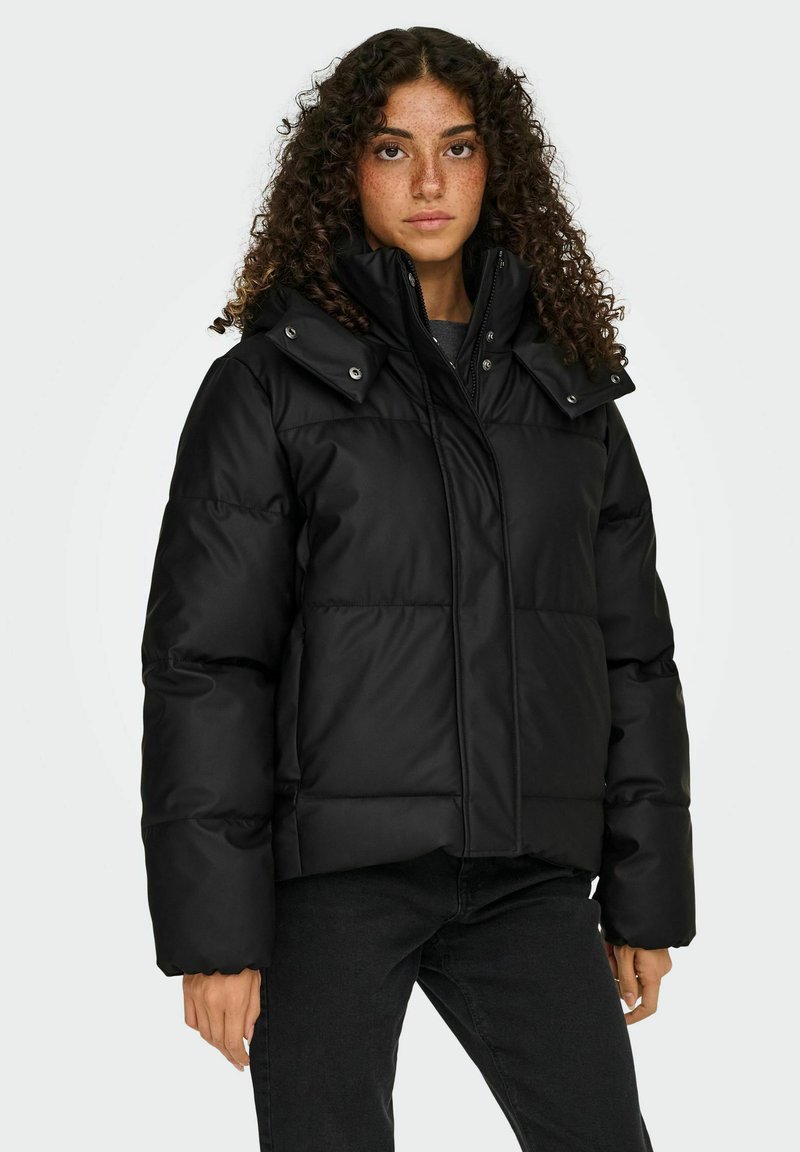 Crna puffer jakna s visokim ovratnikom, kopčama na dugmad i kvadratnim dizajnom. Lagani materijal, oversized kroj, nosi se preko tamne majice.