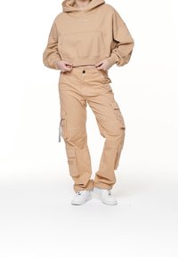 Sudadera corta de color beige con bolsillo frontal, combinada con pantalones cargo beige que cuentan con múltiples bolsillos y una etiqueta lateral, usados con zapatillas blancas.