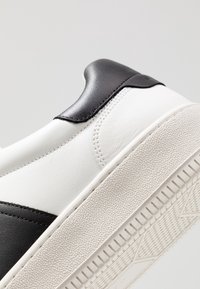 Vit och svart sneaker med en slät läderöverdel, som har en texturerad gummisul, kontrasterande färger och stickade detaljer.