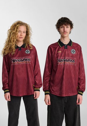 Dva mlada odrasla človeka v ujemajočih se maroon dolgih športnih majicah s logotipi "SWARM INTERNATIONALS", ki stojita ob preprosti beli ozadju.