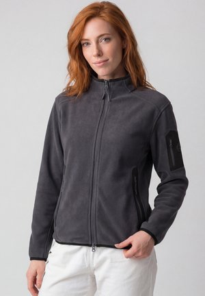 Frau mit roten Haaren, die eine dunkelgraue Fleecejacke mit Reißverschlusstaschen und weiße Hose trägt, steht vor einem schlichten hellen Hintergrund.