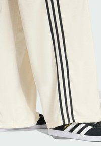 Krémové sportovní kalhoty s třemi černými pruhy po stranách, vyrobené z měkké tkaniny. V kombinaci s černobílými teniskami se zlatými doplňky.
