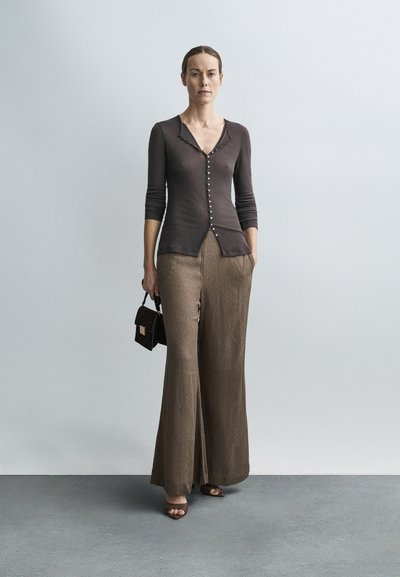 Donna in piedi con maglia marrone abbottonata, pantaloni larghi, sandali, che tiene una piccola borsa a mano, su sfondo semplice grigio chiaro.