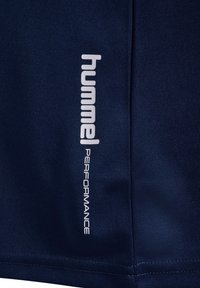 Nahaufnahme eines marineblauen Stoffes mit weißem, vertikalem "hummel PERFORMANCE" Markenlogo, das nahe einer Naht gedruckt ist.