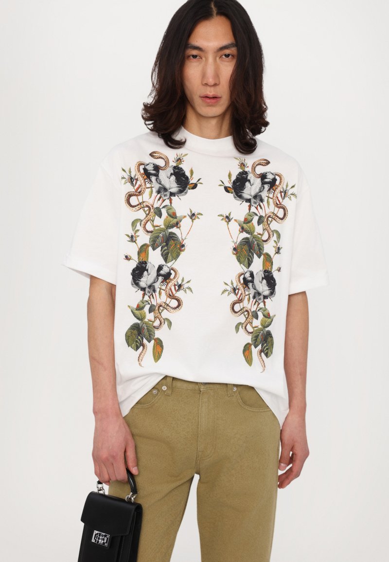 T-shirt oversize bianco con vivaci ricami botanici e animali. Abbinato a pantaloni sabbia con texture e una piccola borsa nera.