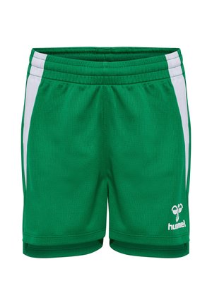 Uśmiechnięta dziewczyna stoi, ubrana w zielony strój sportowy z białymi paskami, białe skarpety do kolan i czarne buty halowe.