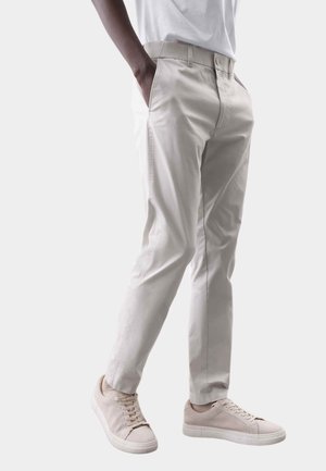Person iført beige chinos med sidelommer og lyse beige lave sneakers, stående med hænderne i lommerne på en hvid baggrund.