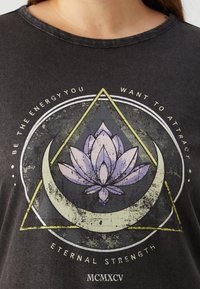 Melnas kokvilnas T-krekls ar nolietotu grafisko dizainu, kurā attēlots lotosu zieds trīsstūrī un pusmēnesī, akcentēts ar baltu un dzeltenu tekstu.