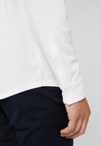White long-sleeve shirt displayed with a buttoned cuff, featuring a smooth texture and straight hem, paired with dark navy pants.
