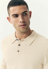 Beige poloko s dlouhým rukávem vyrobené z texturované pleteniny, které má klasický límec a tři tmavé knoflíky na přední léze.