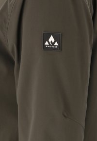 Giacca verde scuro con una texture liscia, dotata di un'etichetta quadrata nera con un logo di montagna bianco e il nome del marchio "WHISTLER."