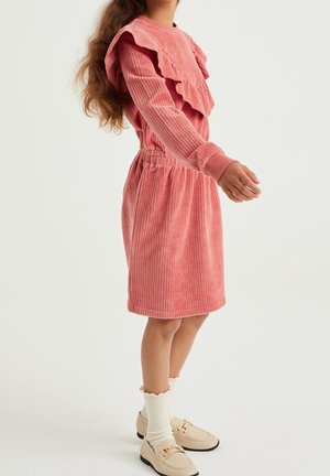 Enfant portant une robe en velours côtelé rose avec des volants, des chaussettes crème et des mocassins beige sur un fond clair uni.