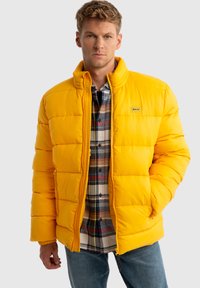 Geltona paminkštinta striukė su aukštu apykakle ir užtrauktuku, turinti horizontalius siūlus ir logotipo pleistrą, dėvima ant languoto flanelinio marškinio.