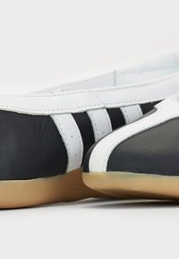 Black and white leather shoes with a smooth texture, featuring a rounded toe and two white accent stripes. Rubber sole. Close-up view.