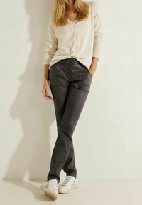 Beige knappar cardigan bärs över mörkgrå tajta byxor med framfickor och sömmar, ihop med vita sneakers med grå detaljer.