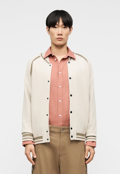 Νεαρός άνδρας που φοράει ένα μπεζ varsity μπουφάν με ριγέ μανσέτες πάνω από ένα ροδακινί πουκάμισο με κουμπιά και μπεζ παντελόνι, στεκόμενος μπροστά σε μια απλή φόντο.