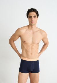 Marineblaue Stretch-Baumwoll-Boxershorts mit einem weichen, pfirsichfarbenen Bund, der wiederholt mit "GANT" branding versehen ist. Das Modell zeigt ein figurbetontes Design.