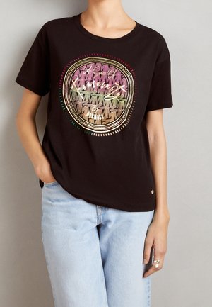 Osoba ubrana w czarną koszulkę z kolorową, okrągłą grafiką oraz jasnoniebieskie dżinsy, z ręką w kieszeni, na tle jednolitej, szarej ściany.