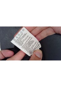 Štítek s pokyny pro údržbu na látce. Bílá látka s černým textem uvádějící pokyny pro údržbu a symboly praní. Držena v ruce proti tmavému pozadí.