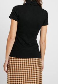 Černý žebrovaný top s krátkým rukávem a rolákem, spárovaný s sukní s houndstooth vzorem v béžové a hnědé barvě. Důraz na texturu a střih.