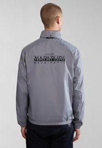 Graue Windjacke mit hohem Kragen, elastischen Bündchen und Saum. Merkmale ein auffälliges schwarzes Logo auf der Rückseite. Leicht und mit glatter Textur.