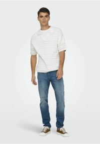 Hvit strikket polo genser med korte ermer, båret over en hvit T-skjorte, paret med blå jeans og brune joggesko.