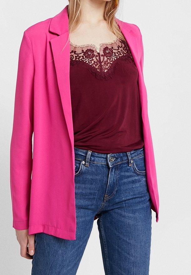 Femme portant un blazer rose vif sur un caraco bordeaux orné de dentelle et un jean bleu, debout devant un fond clair uni.