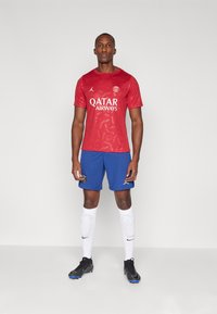 Punane särk, millel on peen logodesain ja valged "QATAR Airways" tekst, koos siniste lühikeste pükste ja mustade jalgpallijalatsitega sportliku ilme saavutamiseks.