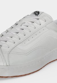 Baskets Vans blanches avec lacets blancs, détail de la bande latérale, couture visible et semelle en caoutchouc marron.
