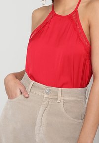 Top rosso a collo halter con dettagli in pizzo, abbinato a pantaloncini in velluto a coste beige con chiusura a bottoni e tasche laterali. Tessuto dalla trama liscia.