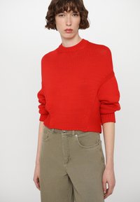 Red knitted sweater with a high neckline, featuring textured patterns and oversized sleeves, paired with light olive green trousers.