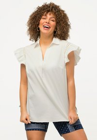 Blouse blanche à manches courtes avec des épaules volantées, un col pointu et un détail en découpe au niveau des coutures des épaules, assortie à un short bleu marine à motifs.