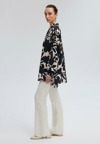 Sort og beige mønstret oversized skjorte med lange ærmer, parret med tætsiddende hvide flared bukser og lyse sko.